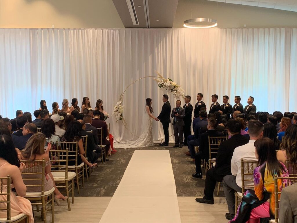 indoor wedding ceremony at brannon center florida with large wall of white curtains and floral arch behind couple